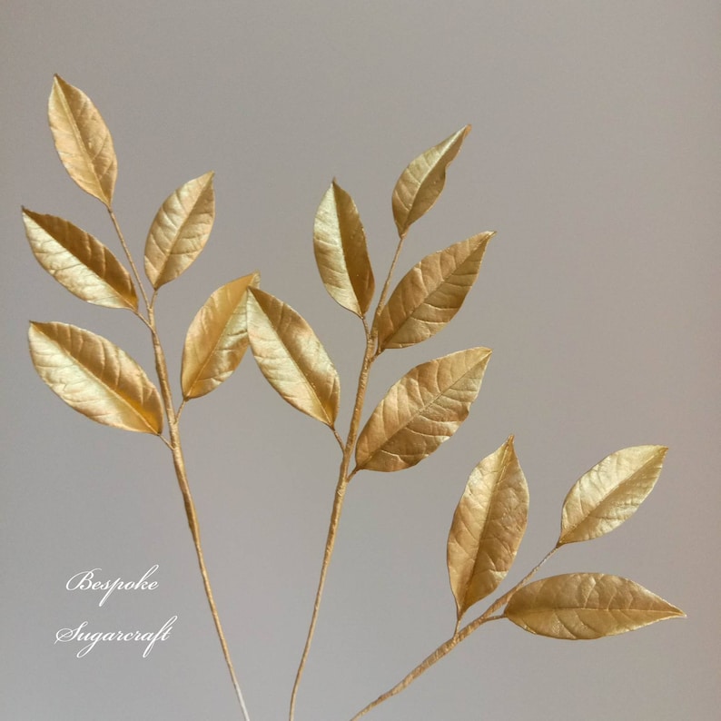 Three gold sugar leaf sprigs with multiple leaves on each stem. The leaves are a metallic gold colour and have a slightly textured surface. The stems are thin and brown. The image has the text "Bespoke Sugarcraft" at the bottom.