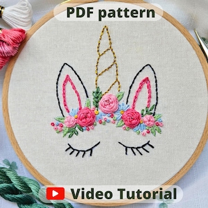 May include: Embroidered unicorn design on a white fabric with pink and blue flowers, a gold horn, and closed eyes. The fabric is stretched over a wooden embroidery hoop. The text "PDF pattern" and "Video Tutorial" are also visible.