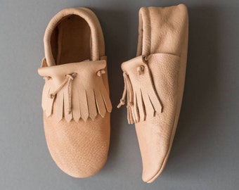 May include: A pair of light brown leather moccasins with fringe detailing. The moccasins have a rounded toe and a simple design.