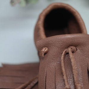 May include: Close-up of a brown leather moccasin with fringe detailing and a tie closure. The moccasin is made of soft, supple leather and has a comfortable fit.