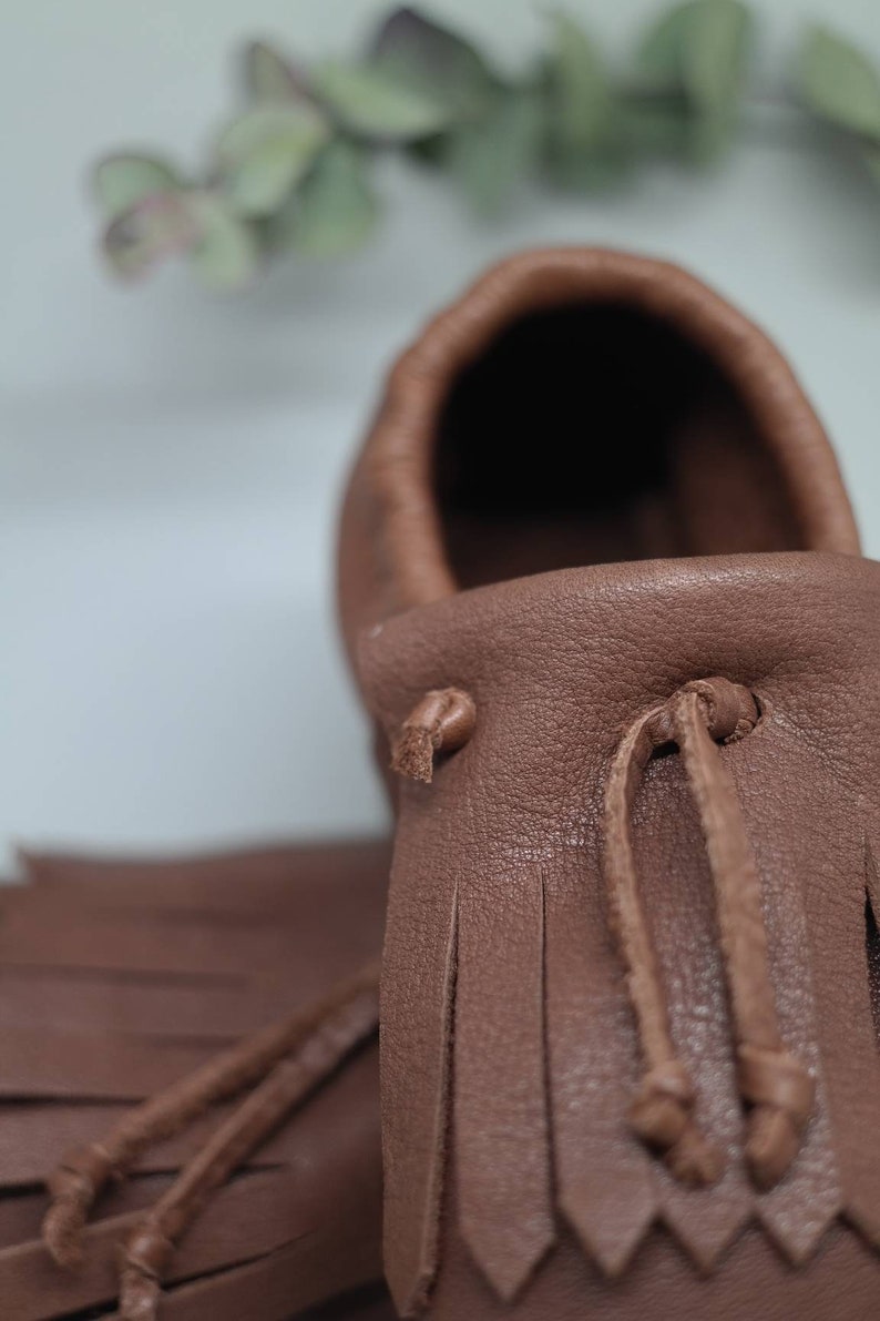 May include: Close-up of a brown leather moccasin with fringe detailing and a tie closure. The moccasin is made of soft, supple leather and has a comfortable fit.