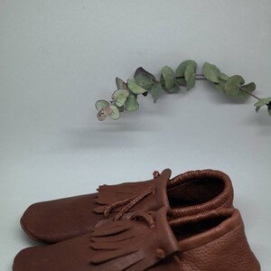 May include: A pair of brown leather moccasins with fringe detailing. The moccasins have a simple design and are made of soft, supple leather.