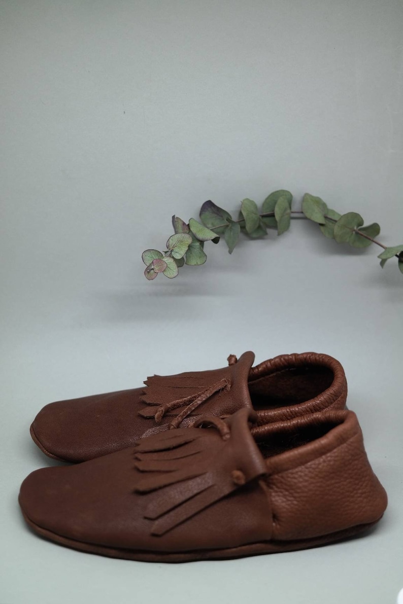 May include: A pair of brown leather moccasins with fringe detailing. The moccasins have a simple design and are made of soft, supple leather.