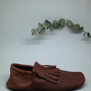 May include: A brown leather moccasin with a fringe detail. The moccasin has a pointed toe and a flat sole.