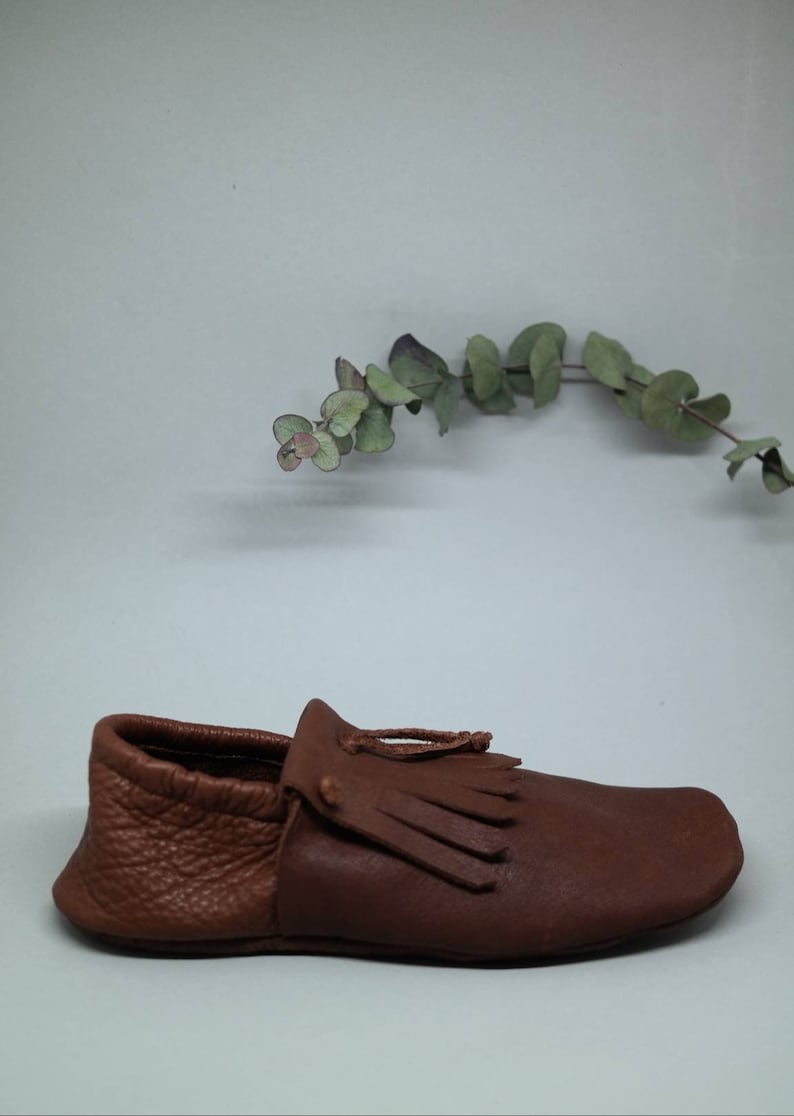May include: A brown leather moccasin with a fringe detail. The moccasin has a pointed toe and a flat sole.