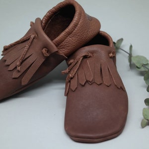 May include: A pair of brown leather moccasins with fringe detailing. The moccasins are soft and comfortable, perfect for babies and toddlers.