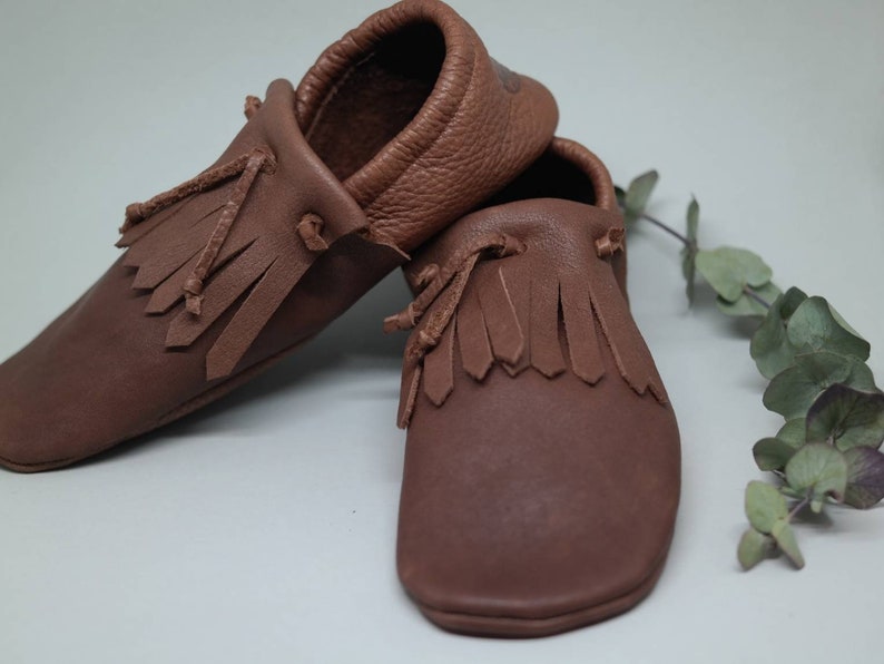May include: A pair of brown leather moccasins with fringe detailing. The moccasins are soft and comfortable, perfect for babies and toddlers.