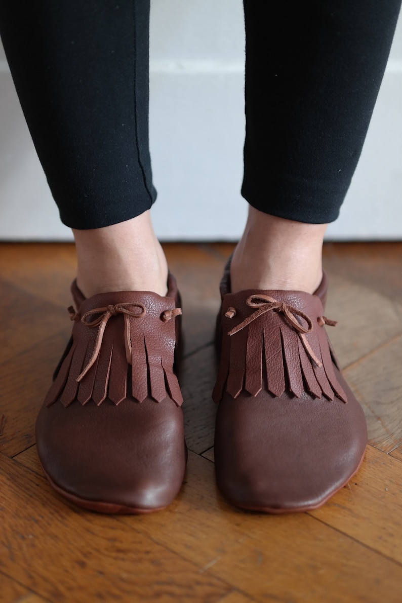 May include: Brown leather shoes with fringe detailing and a bow tie closure.