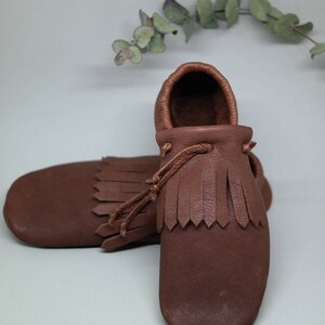 May include: A pair of brown leather moccasins with fringe detailing. The moccasins have a simple design and are made of soft, supple leather.