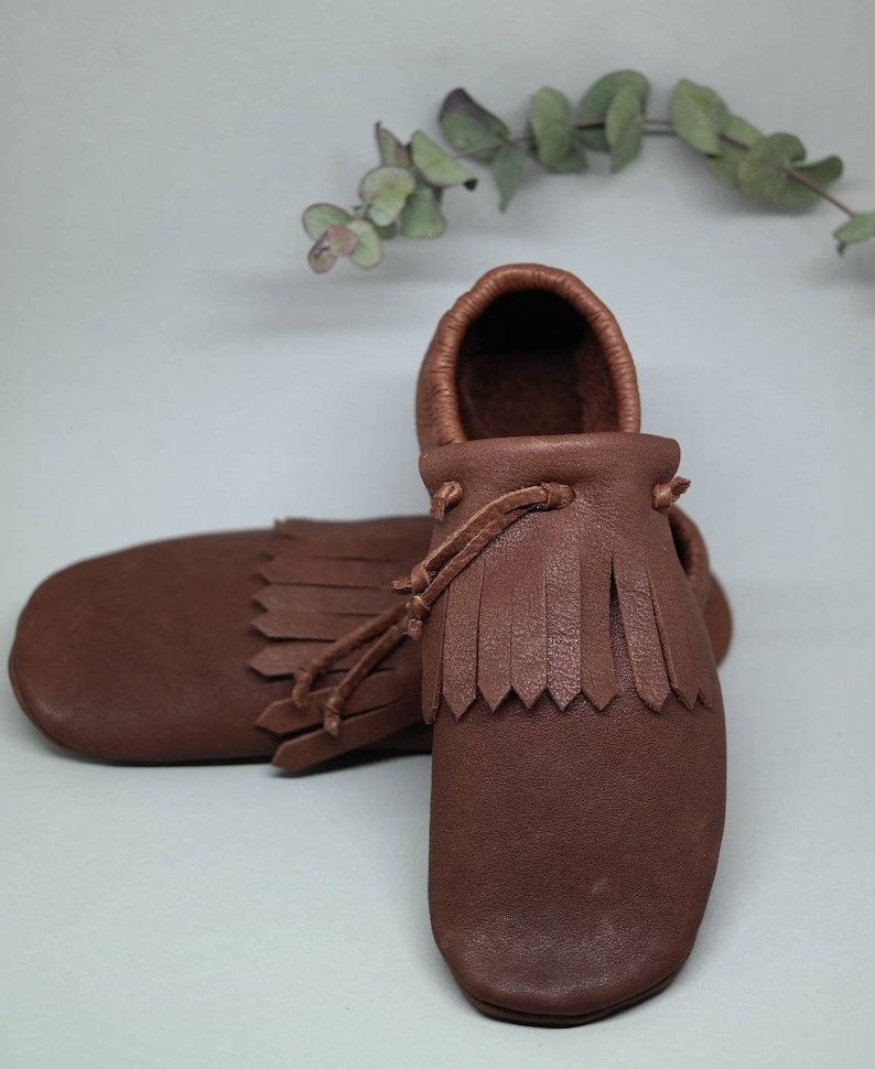 May include: A pair of brown leather moccasins with fringe detailing. The moccasins have a simple design and are made of soft, supple leather.