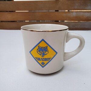 Puede incluir: Taza de cer&aacute;mica color crema con borde dorado y asa curva. La taza presenta un emblema azul y amarillo con la cabeza de un lobo y las palabras "Cub Scouts". Una flor de lis azul est&aacute; debajo del texto.