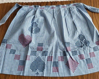 Op de afbeelding: Een blauwe en witte gingham schort met rode geborduurde harten en bloemmotieven. Het schort heeft een geplooide taille en knoopt aan de zijkanten.