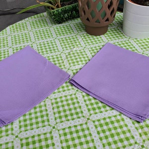 May include: Two lavender-colored cloth napkins are neatly folded and placed on a green and white checkered tablecloth. The napkins are square and appear to be made of a smooth fabric. The tablecloth has a decorative lace-like border.