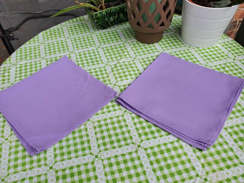 May include: Two lavender-colored cloth napkins are neatly folded and placed on a green and white checkered tablecloth. The napkins are square and appear to be made of a smooth fabric. The tablecloth has a decorative lace-like border.