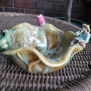 May include: A ceramic bowl with a green frog, a pink flower, and a blue figurine of a person wearing a hat. The bowl is glazed in shades of green, brown, and yellow.