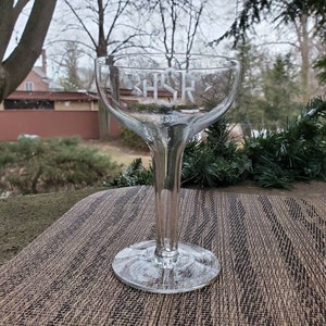 Puede incluir: Una copa de champán de cristal transparente con un monograma "HSR" grabado en blanco en el lateral.