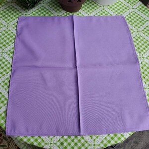 May include: A square, lavender-colored cloth napkin, neatly folded. The napkin is made of a smooth fabric and is displayed on a green and white checkered tablecloth. The napkin is a solid color with a simple design.