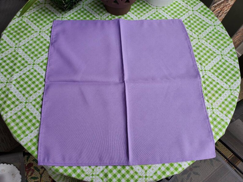 May include: A square, lavender-colored cloth napkin, neatly folded. The napkin is made of a smooth fabric and is displayed on a green and white checkered tablecloth. The napkin is a solid color with a simple design.