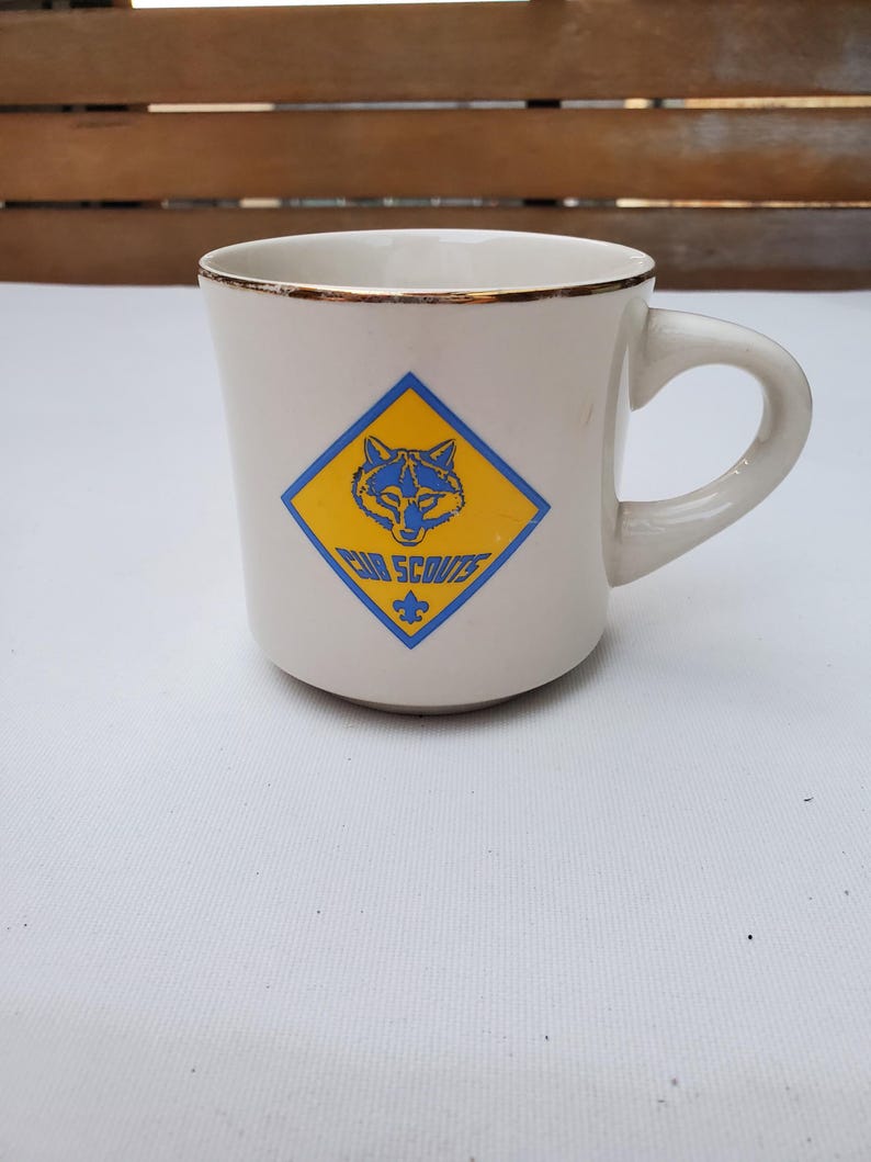 Puede incluir: Taza de cer&aacute;mica color crema con borde dorado y asa curva. La taza presenta un emblema azul y amarillo con la cabeza de un lobo y las palabras "Cub Scouts". Una flor de lis azul est&aacute; debajo del texto.