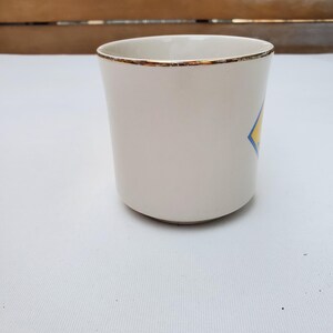 Puede incluir: Una taza de cer&aacute;mica blanca con borde dorado y un dise&ntilde;o en forma de diamante azul y amarillo. La taza tiene una forma cil&iacute;ndrica simple y est&aacute; vac&iacute;a. Est&aacute; sobre una superficie blanca.