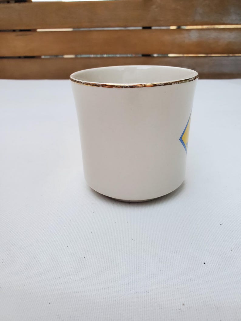 Puede incluir: Una taza de cer&aacute;mica blanca con borde dorado y un dise&ntilde;o en forma de diamante azul y amarillo. La taza tiene una forma cil&iacute;ndrica simple y est&aacute; vac&iacute;a. Est&aacute; sobre una superficie blanca.