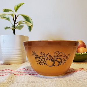 May include: A brown ceramic mixing bowl with a grape and apple design. The bowl is sitting on a white and red tablecloth.