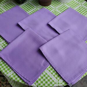 May include: Five square, lavender-colored cloth napkins are arranged on a green and white checkered tablecloth. The napkins are neatly folded, showcasing a smooth, solid color. The image is taken from a slightly elevated angle, providing a clear view of the napkins.