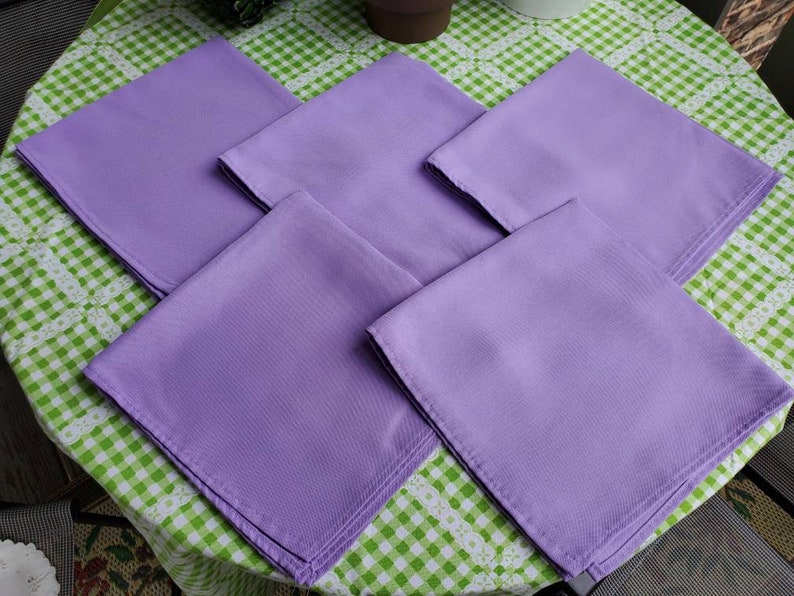 May include: Five square, lavender-colored cloth napkins are arranged on a green and white checkered tablecloth. The napkins are neatly folded, showcasing a smooth, solid color. The image is taken from a slightly elevated angle, providing a clear view of the napkins.