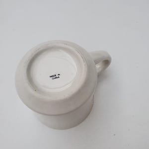 Puede incluir: Una taza de cer&aacute;mica blanca con asa, vista desde abajo. La base de la taza tiene el texto "MADE IN CHINA". La taza es de color liso y neutro, adecuada para diversas bebidas.