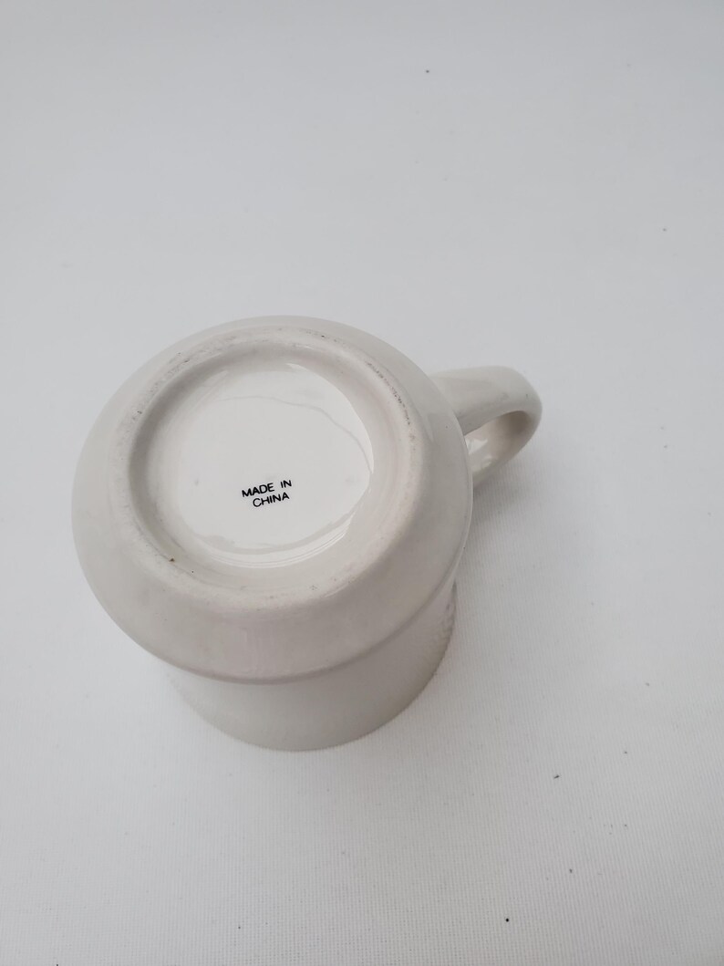 Puede incluir: Una taza de cer&aacute;mica blanca con asa, vista desde abajo. La base de la taza tiene el texto "MADE IN CHINA". La taza es de color liso y neutro, adecuada para diversas bebidas.