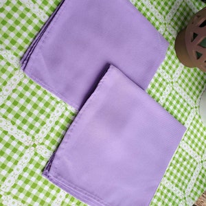 May include: Two folded, square lavender napkins. The napkins are made of a smooth fabric and are neatly stacked. They are resting on a green and white checkered tablecloth with a white lace trim.