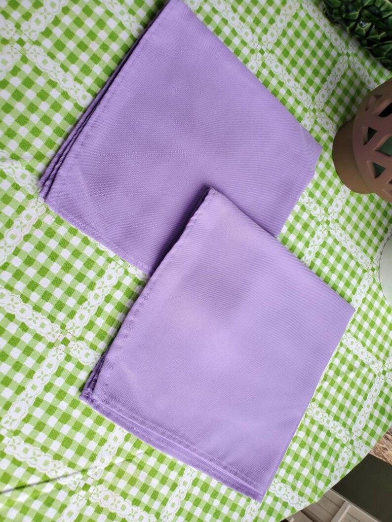 May include: Two folded, square lavender napkins. The napkins are made of a smooth fabric and are neatly stacked. They are resting on a green and white checkered tablecloth with a white lace trim.