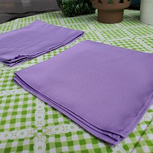 May include: Two lavender cloth napkins are neatly folded and stacked on a green and white checkered tablecloth. The napkins are made of a smooth fabric and are a solid color. The tablecloth has a decorative white lace trim.