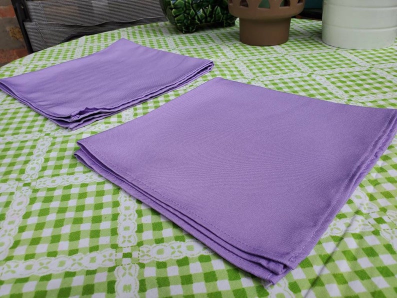 May include: Two lavender cloth napkins are neatly folded and stacked on a green and white checkered tablecloth. The napkins are made of a smooth fabric and are a solid color. The tablecloth has a decorative white lace trim.