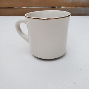 Puede incluir: Una taza de cer&aacute;mica blanca con un borde dorado y un asa curvada. La taza tiene un dise&ntilde;o simple y elegante y est&aacute; colocada sobre una superficie blanca y un fondo de madera.