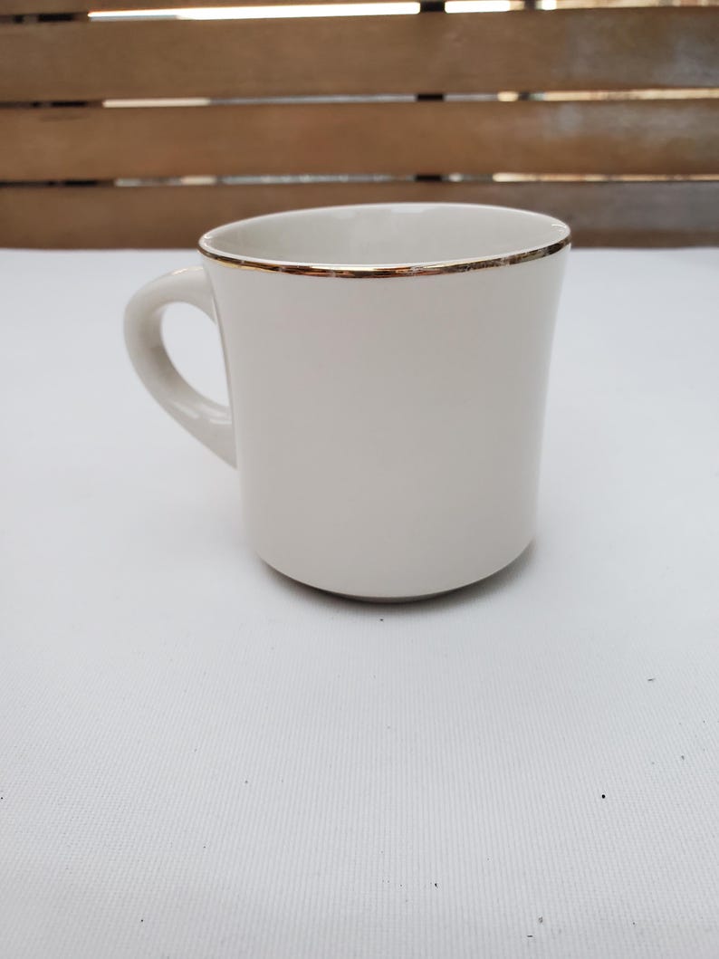 Puede incluir: Una taza de cer&aacute;mica blanca con un borde dorado y un asa curvada. La taza tiene un dise&ntilde;o simple y elegante y est&aacute; colocada sobre una superficie blanca y un fondo de madera.