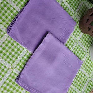 May include: Two lavender-colored cloth napkins are neatly folded and arranged on a green and white checkered tablecloth. The napkins have a smooth texture and are positioned diagonally, showcasing their rectangular shape.