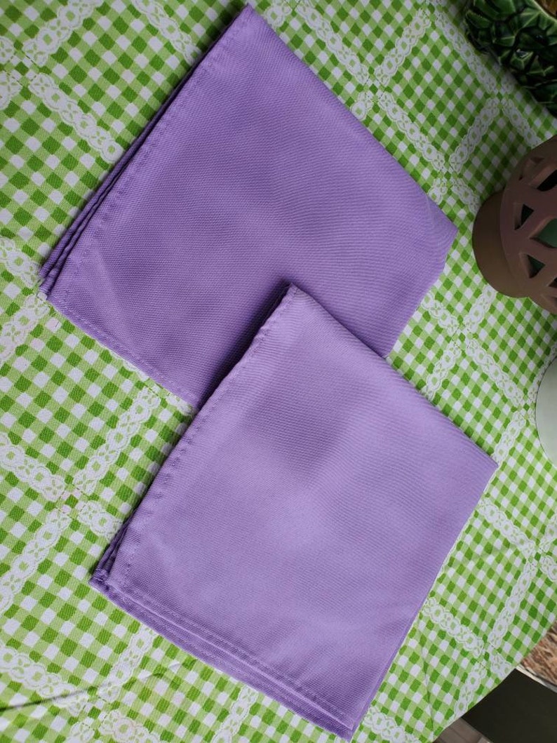 May include: Two lavender-colored cloth napkins are neatly folded and arranged on a green and white checkered tablecloth. The napkins have a smooth texture and are positioned diagonally, showcasing their rectangular shape.