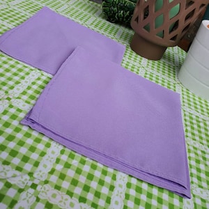 May include: Two folded lavender napkins are placed on a green and white checkered tablecloth. The napkins are a solid color and appear to be made of a smooth fabric. The tablecloth has a decorative white lace trim.