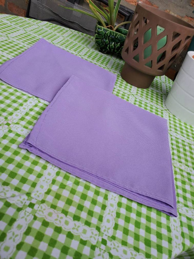 May include: Two folded lavender napkins are placed on a green and white checkered tablecloth. The napkins are a solid color and appear to be made of a smooth fabric. The tablecloth has a decorative white lace trim.