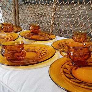Puede incluir: Juego de seis platos de postre y tazas de vidrio ámbar. Los platos tienen un borde festoneado y un patrón en relieve. Las tazas tienen un patrón similar y una forma redondeada.