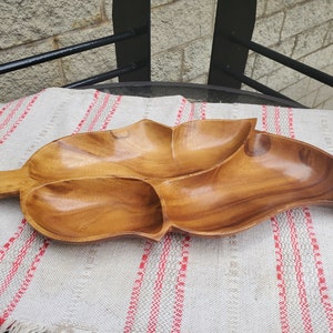 Puede incluir: Una bandeja de madera con forma de hoja con tres compartimentos. La bandeja es de madera de color marrón claro y tiene un acabado liso y pulido. Descansa sobre un paño a rayas blancas y rojas.