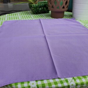 May include: A square, solid lavender-colored cloth napkin. The napkin is made of a smooth fabric and is neatly folded. It is placed on a green and white checkered tablecloth. The napkin is a simple, elegant piece.