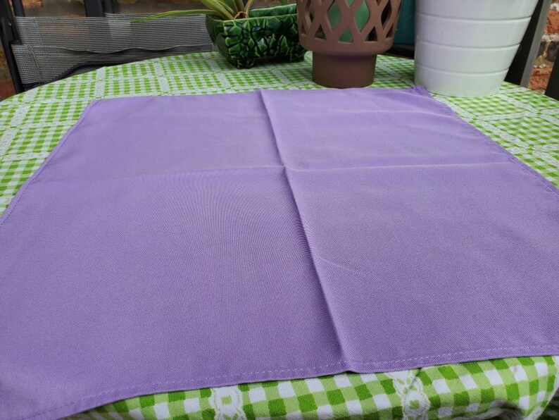 May include: A square, solid lavender-colored cloth napkin. The napkin is made of a smooth fabric and is neatly folded. It is placed on a green and white checkered tablecloth. The napkin is a simple, elegant piece.