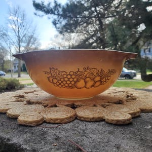 Peut inclure: Un bol mélangeur Pyrex brun et blanc avec un motif de fruits. Le bol est posé sur un tapis de paille tissé.