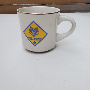 Puede incluir: Taza de cer&aacute;mica blanca con borde dorado y asa curva. La taza presenta un rombo azul y amarillo con un lobo y las palabras "Cub Scouts". La taza est&aacute; sobre una superficie blanca.
