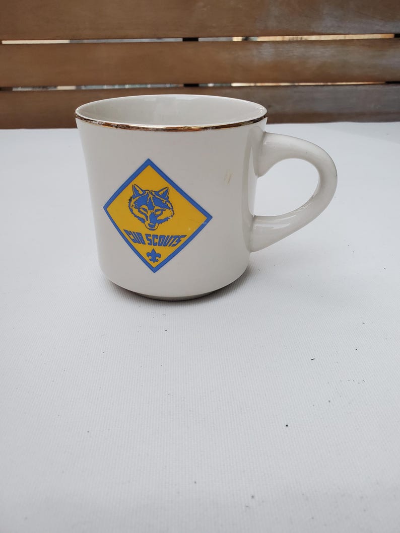 Puede incluir: Taza de cer&aacute;mica blanca con borde dorado y asa curva. La taza presenta un rombo azul y amarillo con un lobo y las palabras "Cub Scouts". La taza est&aacute; sobre una superficie blanca.