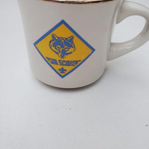 Puede incluir: Taza de cer&aacute;mica blanca con borde dorado y asa curva. La taza presenta un logotipo de los Cub Scouts azul y amarillo con la cabeza de un lobo y las palabras "Cub Scouts".
