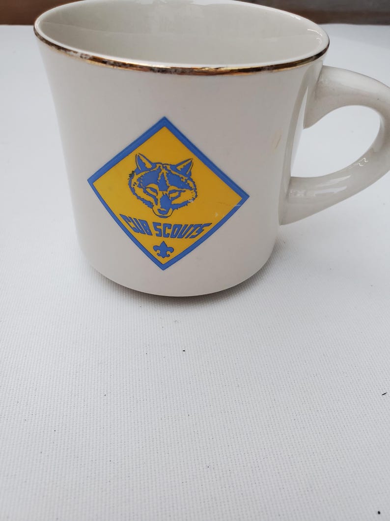 Puede incluir: Taza de cer&aacute;mica blanca con borde dorado y asa curva. La taza presenta un logotipo de los Cub Scouts azul y amarillo con la cabeza de un lobo y las palabras "Cub Scouts".
