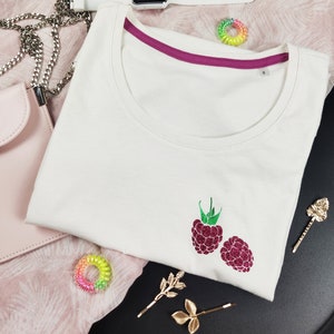 May include: A white t-shirt with a scoop neck and a raspberry design. The shirt has a magenta trim on the neckline and a small size tag. Accessories include a pink purse, a white smartwatch, and gold hair clips.